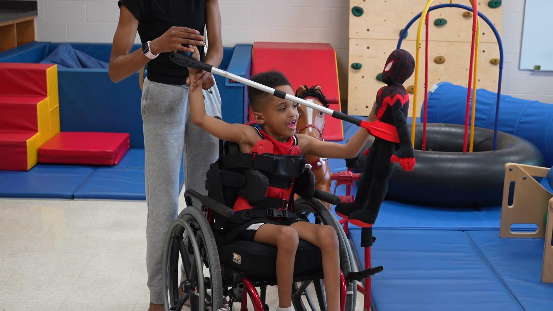 A boy in a wheelchair using a 3D printed grabber to pick up a spiderman toy.